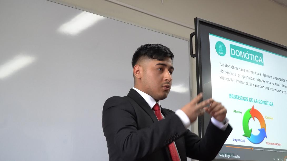 Estudiante vestido formal, con chaqueta oscura y corbata roja expone ante la comusión con una pantalla de fondo que muestra su tesis